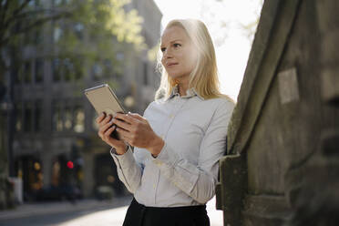 Thoughtful blond businesswoman looking away while holding digital tablet and leaning on surrounding wall in city - JOSEF00988