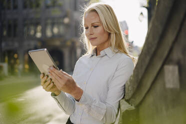 Blonde, hübsche Unternehmerin, die ein digitales Tablet benutzt, während sie sich in der Stadt an eine Wand lehnt - JOSEF00990