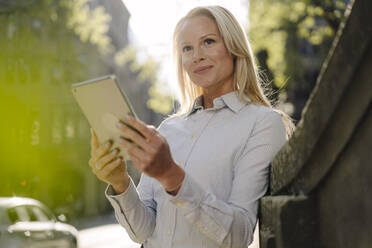 Smiling blond female entrepreneur looking away while leaning on surrounding wall with digital tablet in city - JOSEF00992