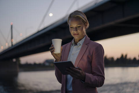 Beautiful businesswoman using digital tablet while holding disposable cup against bridge in city at dusk stock photo