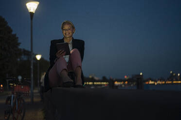 Smiling beautiful female entrepreneur using digital tablet while sitting on retaining wall against sky at night - JOSEF00994