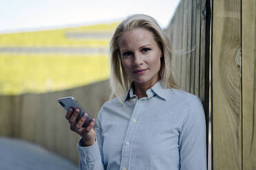 Confident blond businesswoman holding smart phone against wooden surrounding wall - JOSEF01009