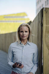 Confident beautiful blond businesswoman holding smart phone against wooden surrounding wall - JOSEF01010