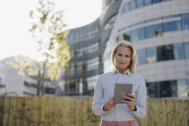 Confident businesswoman holding digital tablet while standing against building at financial district - JOSEF01018