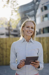 Smiling blond businesswoman holding digital tablet at financial district in city - JOSEF01022