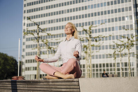 Geschäftsfrau beim Meditieren im Lotussitz auf einer Bank vor einem Gebäude an einem sonnigen Tag, lizenzfreies Stockfoto