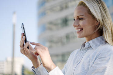 Nahaufnahme einer lächelnden blonden Geschäftsfrau, die ein Smartphone im Finanzviertel benutzt - JOSEF01043