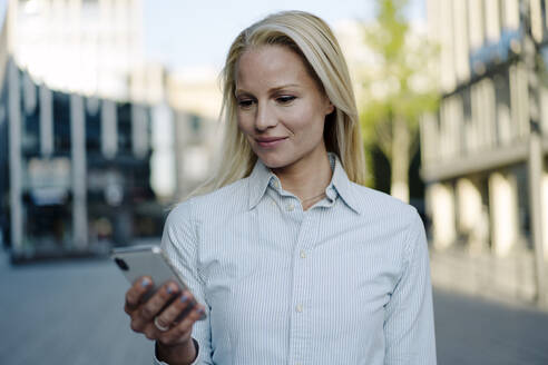 Lächelnde blonde Geschäftsfrau mit Smartphone im Finanzviertel - JOSEF01076