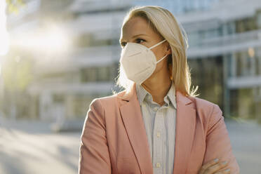 Blond female entrepreneur wearing protective face mask while looking away in financial district during coronavirus - JOSEF01078