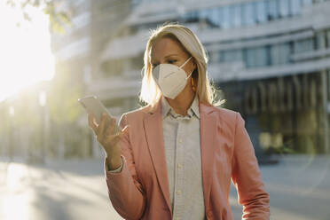 Businesswoman wearing face mask while using smart phone in financial district during pandemic - JOSEF01079