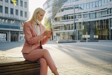 Female professional using smart phone while sitting on bench at financial during coronavirus - JOSEF01083