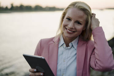 Nahaufnahme einer lächelnden schönen blonden Frau, die bei Sonnenuntergang ein digitales Tablet hält - JOSEF01092