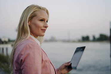 Lächelnde blonde Geschäftsfrau, die ein digitales Tablet hält und bei Sonnenuntergang wegschaut - JOSEF01094