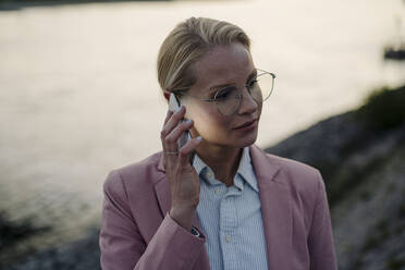 Nahaufnahme einer schönen Geschäftsfrau, die bei Sonnenuntergang am Fluss mit ihrem Mobiltelefon spricht - JOSEF01100