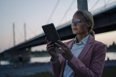 Selbstbewusste Unternehmerin vor einer Brücke in der Abenddämmerung mit digitalem Tablet - JOSEF01105