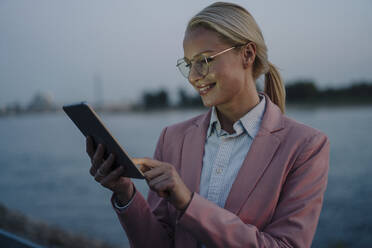 Lächelnde schöne blonde Geschäftsfrau mit digitalen Tablet gegen Fluss in der Abenddämmerung - JOSEF01106