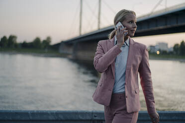 Weibliche Fachkraft, die wegschaut, während sie mit ihrem Smartphone vor einer Brücke in der Abenddämmerung spricht - JOSEF01110