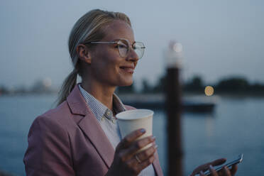 Smiling female entrepreneur holding disposable cup and mobile phone while looking away at dusk - JOSEF01111