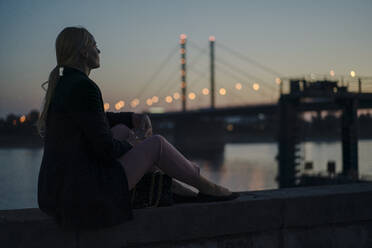 Female professional sitting on retaining wall by river at dusk - JOSEF01113