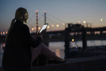 Weibliche Fachkraft, die ein digitales Tablet benutzt, während sie in der Abenddämmerung auf einer Stützmauer sitzt - JOSEF01116