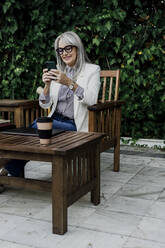 Smiling senior woman sitting on chair while using smart phone at garden - ERRF04068