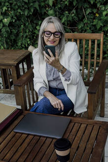 Lächelnde Unternehmerin, die ein Smartphone benutzt, während sie mit einem Laptop am Tisch im Garten sitzt - ERRF04069
