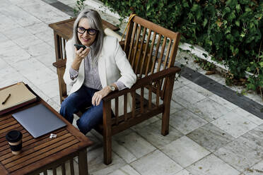 Lächelnde ältere Geschäftsfrau, die über einen Smartphone-Lautsprecher spricht, während sie im Garten sitzt - ERRF04071