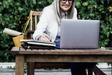 Lächelnde ältere Geschäftsfrau, die im Garten am Laptop in ihr Tagebuch schreibt - ERRF04078