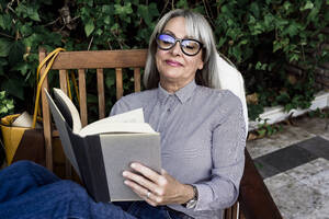 Relaxed female professional reading book while sitting on chair at garden - ERRF04094