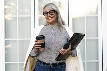 Smiling senior businesswoman holding disposable cup with file and smart phone against office door - ERRF04101