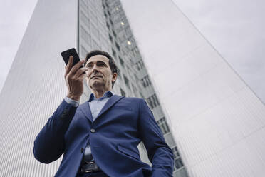 Mature businessman with smartphone in front of an office tower in the city - JOSEF01146