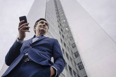 Mature businessman with smartphone in front of an office tower in the city - JOSEF01147