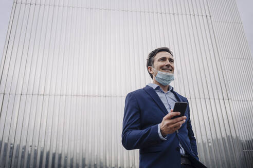 Smiling mature businessman with protective face mask and smartphone in the city - JOSEF01148