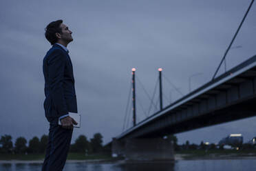 Mature businessman holding tablet standing at the riverbank at dusk - JOSEF01167