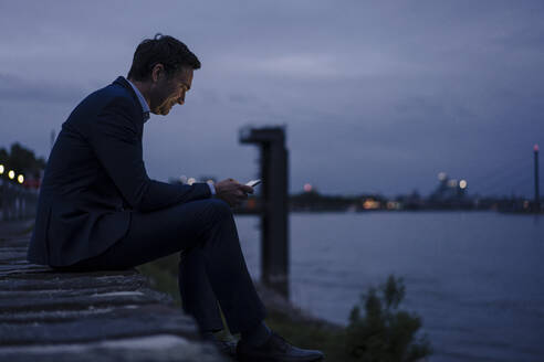 Mature businessman using tablet at the riverbank at dusk - JOSEF01168