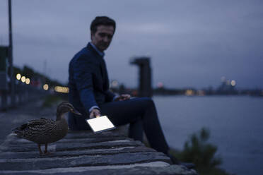 Älterer Geschäftsmann mit Ente und Tablet am Flussufer in der Abenddämmerung - JOSEF01169