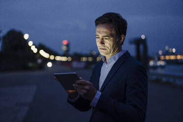 Mature businessman using tablet at the riverbank at dusk - JOSEF01174