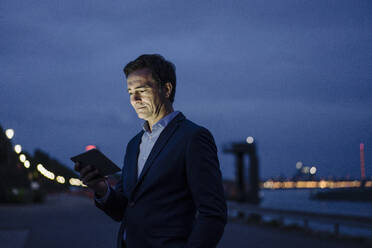 Mature businessman using tablet at the riverbank at dusk - JOSEF01175