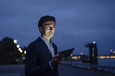 Mature businessman holding tablet at the riverbank at dusk - JOSEF01176