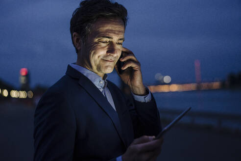 Mature businessman using tablet and smartphone at the riverbank at dusk - JOSEF01177