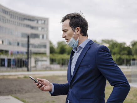 Mature businessman with protective face mask using smartphone in the city - JOSEF01211