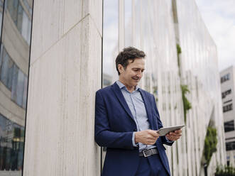 Mature businessman leaning against a building in the city using tablet - JOSEF01214