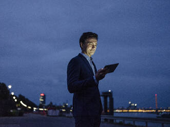 Mature businessman using tablet at the riverbank at dusk - JOSEF01227