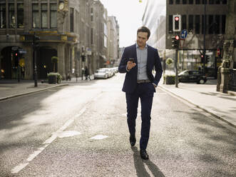Mature businessman walking on a city street using smartphone - JOSEF01236
