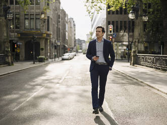 Mature businessman walking on a city street holding smartphone - JOSEF01237