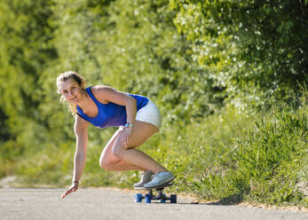 Schöne junge Frau skateboarding auf der Straße von Pflanzen während sonnigen Tag - STSF02554