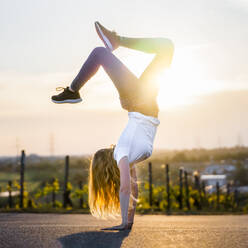 Back lit young woman doing handstand on road - STSF02563