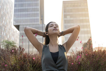 Entspannte junge Geschäftsfrau, die in der Pause mit den Händen hinter dem Kopf vor einem Bürogebäude Musik über Kopfhörer hört - AFVF06705