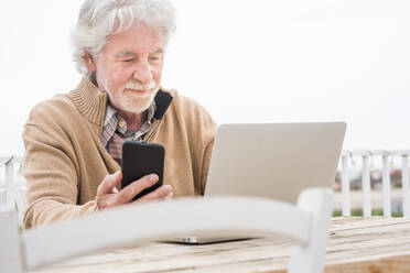 Senior man holding mobile phone while using laptop at table - SIPF02176