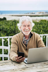 Smiling senior man holding smart phone while sitting with laptop at table - SIPF02177